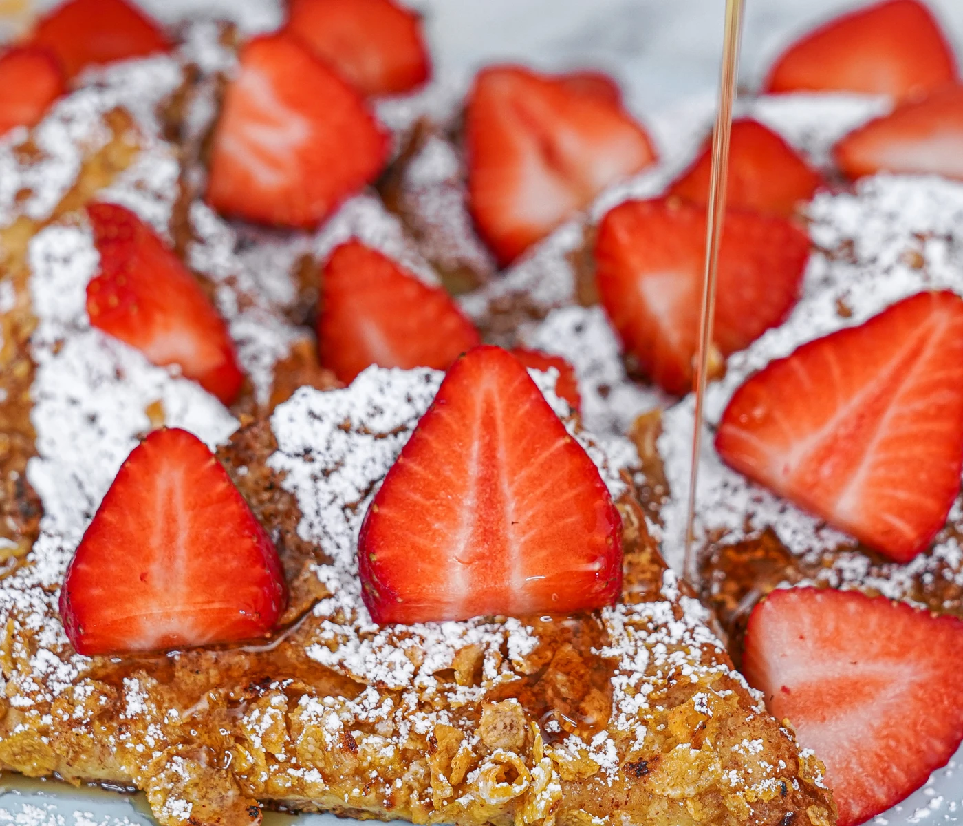 Syrup being poured over crunchy cereal french toast with strawberries.