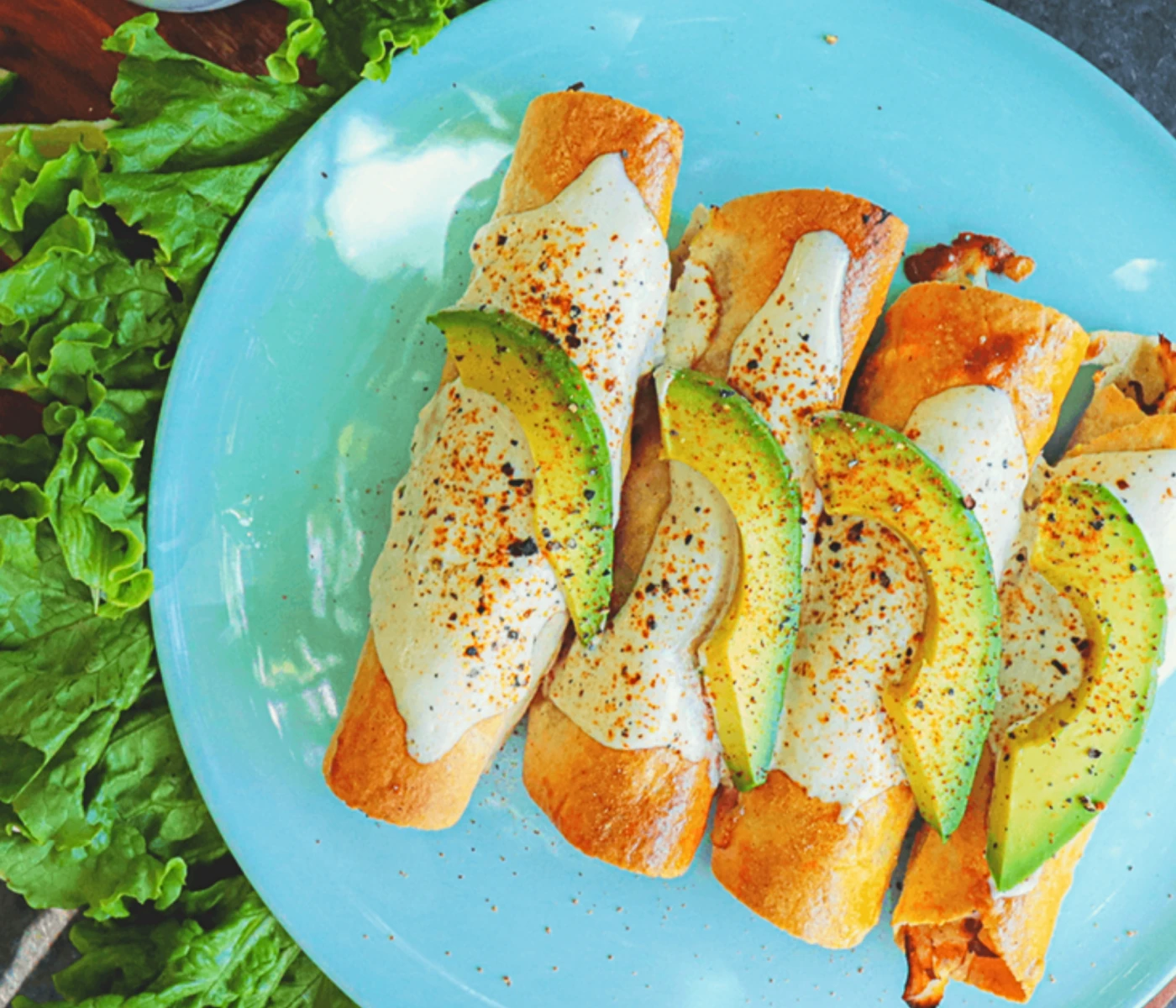 Chicken taquitos topped with a white sauce and avocado.